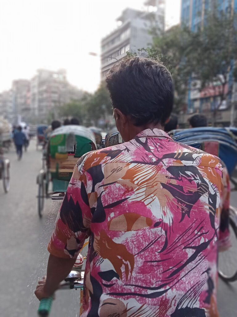 View from a rickshaw passenger seat behind the rickshaw puller on a busy Dhaka street